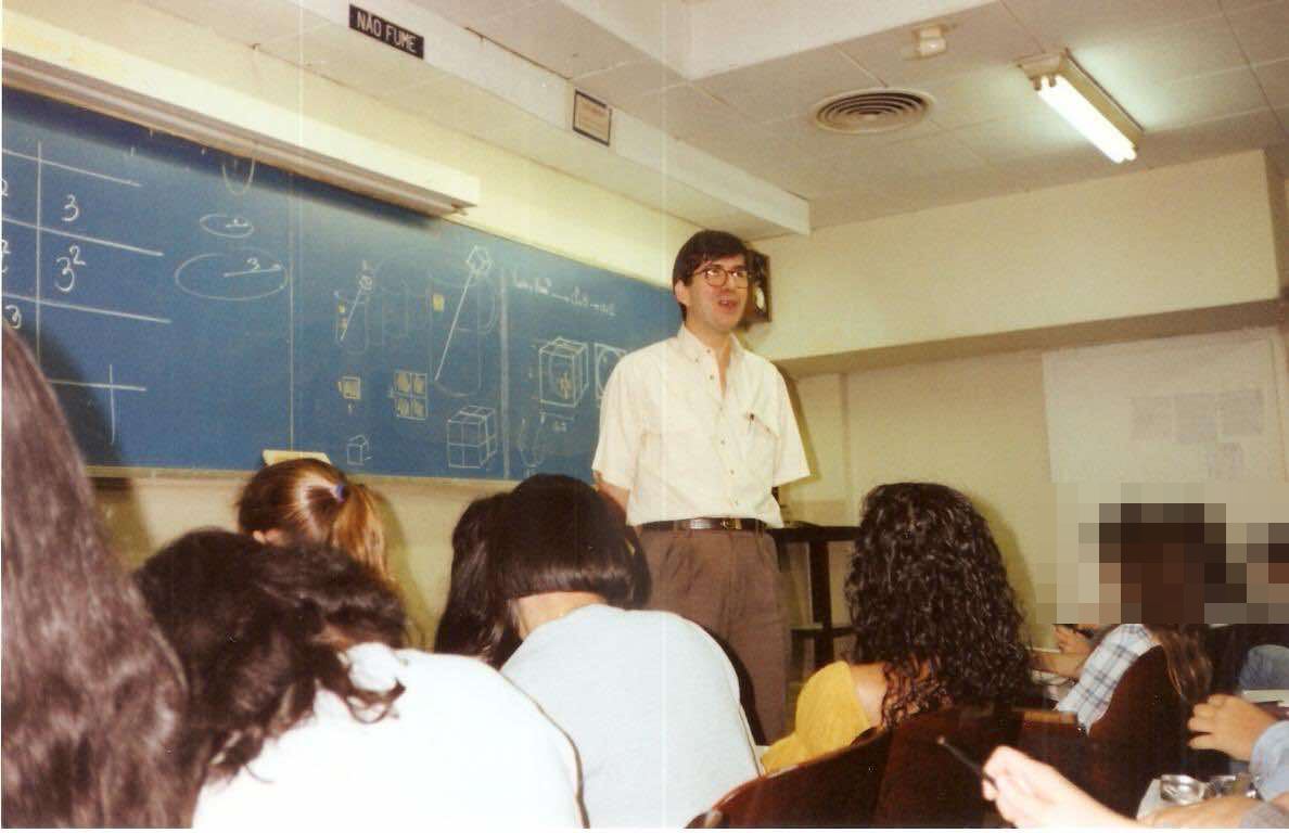 Prof. Anselmo Pisco diante da lousa cheia de desenhos de geometria, falando com a turma